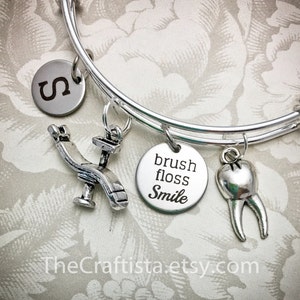 Könnte beinhalten: Silbernes Charm-Armband mit Zahnthema-Anhängern, darunter ein Zahn, ein Zahnarztstuhl und ein Anhänger mit der Aufschrift "brush floss smile".