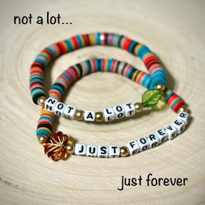 Puede incluir: Dos pulseras de cuentas coloridas con las palabras "not a lot" y "just forever" escritas en letras blancas sobre cuentas negras. Las pulseras están sobre una superficie de madera clara.