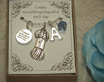 Puede incluir: Un collar de plata con un dije de ovillo de lana, un dije de la letra A y un dije de cristal azul. El collar viene en una pequeña caja con el texto "Crea algo hermoso cada día".