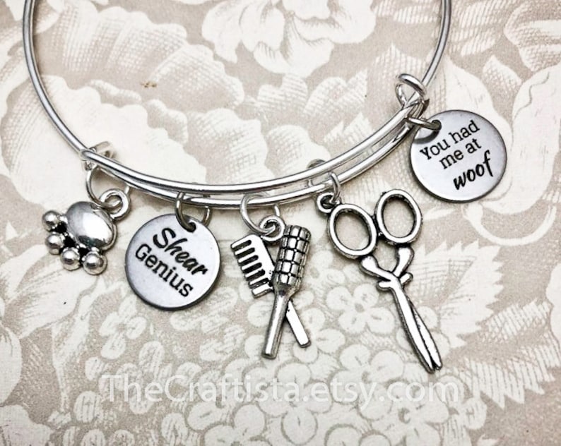May include: Silver charm bracelet with multiple charms, including a paw print, scissors, a comb and brush, and two round charms that say "Shear Genius" and "You had me at woof".