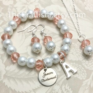 May include: A jewelry set featuring a bracelet, earrings, and a necklace. The bracelet, earrings, and necklace have white faux pearls and pink crystal accents. The bracelet has a silver charm with the letter "A" and a "Junior Bridesmaid" charm.
