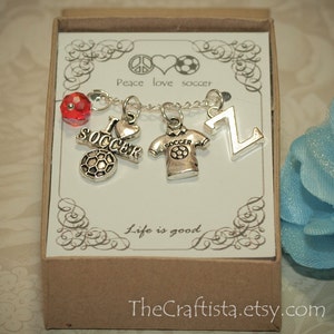 May include: A small gift box with a silver charm bracelet featuring soccer-themed charms, including a soccer ball, a jersey, and the letter Z. The box has a white background with the text "Peace love soccer" and "Life is good".
