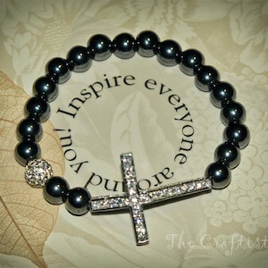 May include: A black beaded bracelet with a silver cross charm and a crystal accent. The bracelet has the words "Inspire you and everyone" written on a white background.