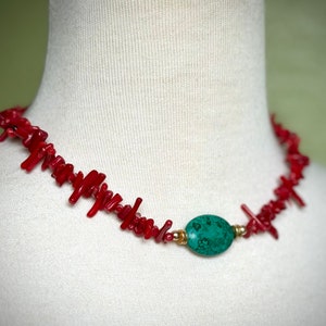 May include: A red coral necklace with a green gemstone pendant. The necklace is made of small, irregular pieces of coral that are strung together on a gold chain. The pendant is a smooth, oval-shaped stone with a green and brown marbled pattern.