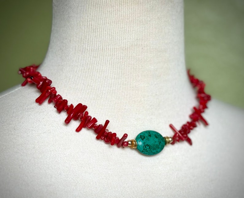 May include: A red coral necklace with a green gemstone pendant. The necklace is made of small, irregular pieces of coral that are strung together on a gold chain. The pendant is a smooth, oval-shaped stone with a green and brown marbled pattern.