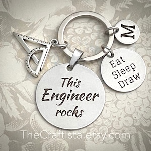 Puede incluir: Llavero de plata con un encanto de transportador, una etiqueta redonda que dice "This Engineer rocks", y otra etiqueta redonda que dice "Eat Sleep Draw".