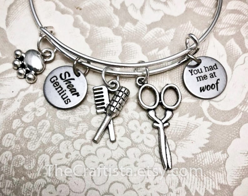 May include: Silver charm bracelet with charms that say "Shear Genius" and "You had me at woof". The bracelet also has charms of a paw print, scissors, a comb, and a brush.