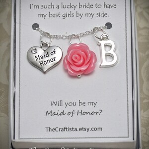 May include: A necklace with a silver chain and charms, including a heart charm with "Maid of Honor", a pink rose, and the letter "B". The card reads "I'm such a lucky bride to have my best girls by my side" and "Will you be my Maid of Honor?"