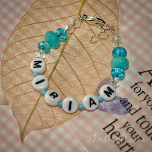 May include: A white beaded bracelet with the name "MIA" spelled out in white letters. The bracelet is made with clear and turquoise blue beads and has a silver clasp.