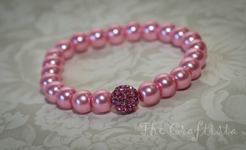K&ouml;nnte beinhalten: Ein rosa Perlenarmband mit einem rosafarbenen Strassstein in der Mitte.