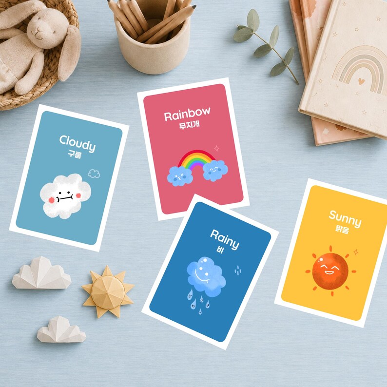 Bilingual weather flashcards (English and Korean) including Cloudy, Rainbow, Rainy, and Sunny, scattered on a blue surface with a bunny plushie and pencils. The cards feature cute hand-drawn weather characters.