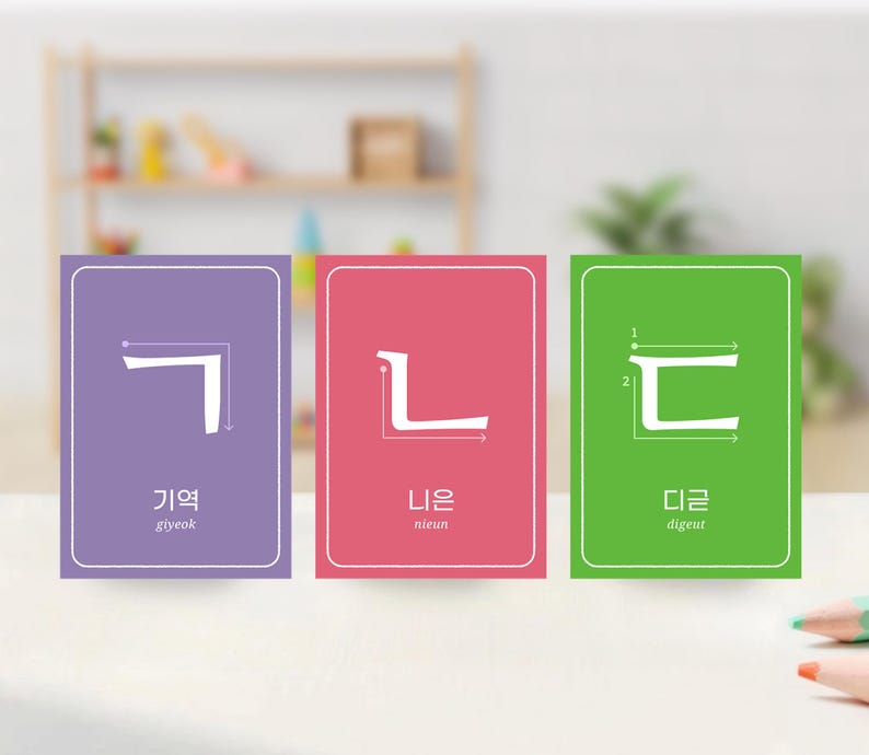 Classroom-style mockup with three large Korean jamo flashcards standing on a table, showing Hangul consonants with stroke order arrows and pronunciation.