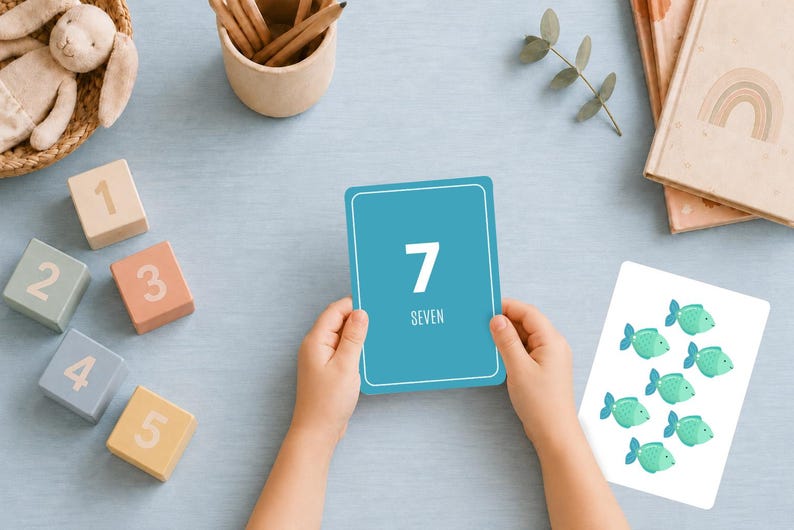 K&ouml;nnte beinhalten: Eine rechteckige blaue Lernkarte mit der wei&szlig;en Zahl 7 und dem Wort SEVEN in Gro&szlig;buchstaben. Daneben zeigt eine wei&szlig;e Karte sieben kleine gr&uuml;ne Fischillustrationen. Die Karten sind von h&ouml;lzernen Zahlenbl&ouml;cken umgeben.