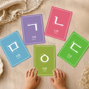 Child’s hands playing with colorful Korean jamo flashcards on a wooden table, showing Hangul consonants with stroke order arrows and English pronunciation.