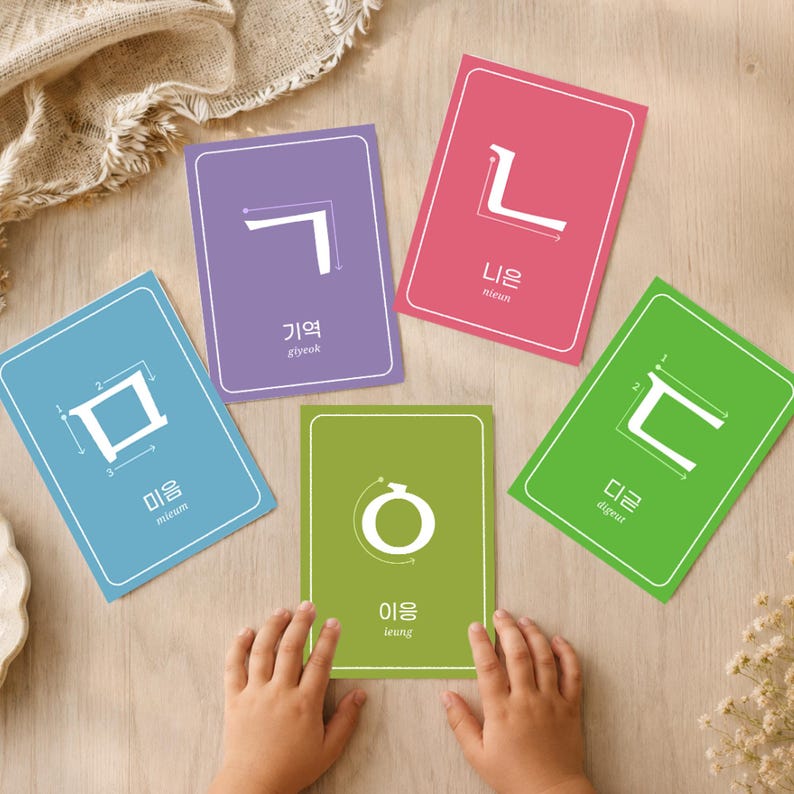 Child’s hands playing with colorful Korean jamo flashcards on a wooden table, showing Hangul consonants with stroke order arrows and English pronunciation.