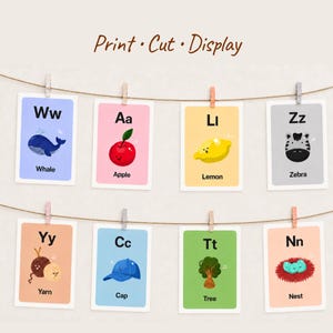 Eight colorful alphabet cards, including Whale, Apple, Lemon, and Zebra, are hung on a string with wooden clothespins against a plain wall. The title 'Print · Cut · Display' suggests home decor or classroom use.