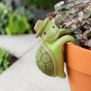 Puede incluir: Una figura de tortuga verde con sombrero, sonriendo y agarrada al borde de una maceta naranja. La tortuga tiene un caparazón detallado y está colocada junto a una planta con hojas rosas y verdes. El fondo está borroso.