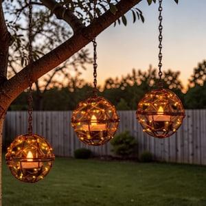 以下が含まれることがあります： 温かい黄色のライトとキャンドルが入った、3つの球形の吊り下げ式ランタン。ランタンは、チェーンで木の枝から吊り下げられています。背景には、緑の芝生、木の柵、夕焼けがあります。