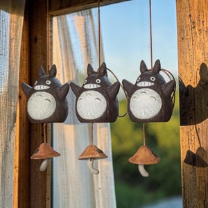 Puede incluir: Tres campanillas decorativas colgantes con un personaje de dibujos animados con cuerpo marrón oscuro, vientre blanco y orejas pequeñas. Cada campanilla tiene un pequeño acento en forma de hongo. Las campanillas están suspendidas por una cuerda marrón.