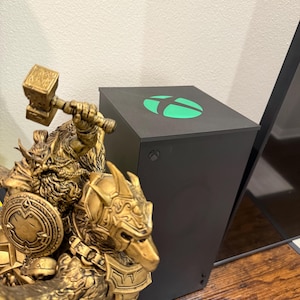 May include: A black Xbox Series X console with a green logo is displayed next to a gold-colored statue of a warrior holding a hammer and shield. The console has a sleek, minimalist design.