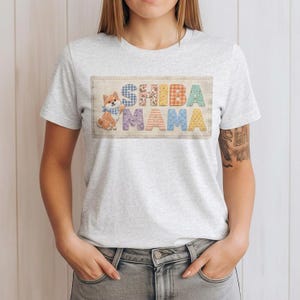 以下が含まれることがあります： 明るい灰色のTシャツ。カラフルなパッチワーク文字で「SHIBA MAMA」と書かれています。青いバンダナをつけた柴犬が左側にいます。このシャツは柔らかい素材で作られています。