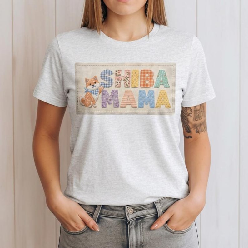 以下が含まれることがあります： 明るい灰色のTシャツ。カラフルなパッチワーク文字で「SHIBA MAMA」と書かれています。青いバンダナをつけた柴犬が左側にいます。このシャツは柔らかい素材で作られています。