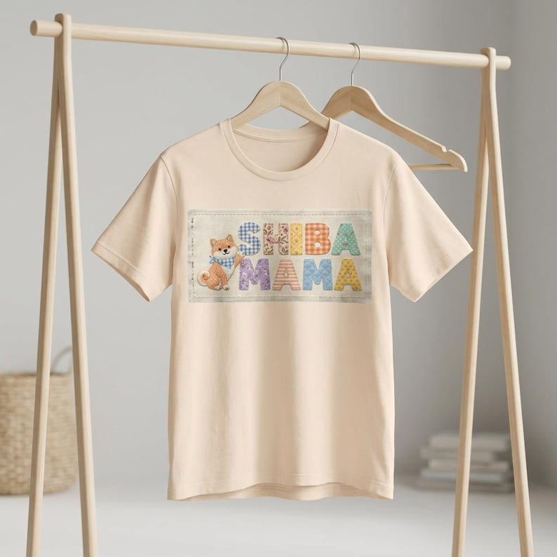以下が含まれることがあります： 明るいピーチ色のTシャツ。カラフルなパッチワーク文字で「SHIBA MAMA」と書かれています。左側には青いスカーフをつけた漫画の柴犬がいます。Tシャツは木製のハンガーラックに吊るされています。