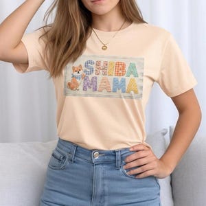 以下が含まれることがあります： 明るいピーチ色のTシャツ。カラフルな模様の文字で「SHIBA MAMA」と書かれています。青いバンダナをつけた漫画の柴犬が左側にいます。柔らかい素材でできた、半袖のTシャツです。