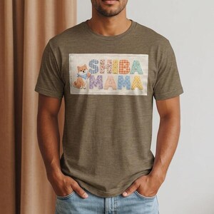 以下が含まれることがあります： カラフルなグラフィックが特徴のヘザーブラウンのTシャツ。グラフィックには、パッチワーク風の文字で「Shiba Mama」という文字と、青いバンダナをつけた漫画の柴犬が描かれています。このシャツは半袖のクルーネックです。