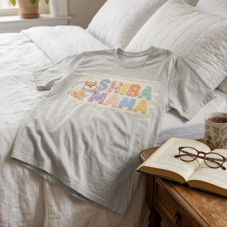 以下が含まれることがあります： 「SHIBA MAMA」の文字がカラフルにプリントされたライトグレーのTシャツ。青いスカーフを巻いた柴犬のパッチが付いています。白い寝具が敷かれたベッドの上にTシャツが置かれています。