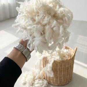 May include: A large pile of fluffy, white material held in a hand, with more spilling into a woven basket. The material appears soft and cloud-like, suggesting a textile or filling material. The basket is light brown.
