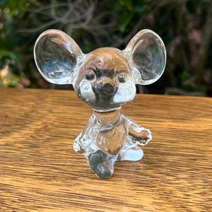 May include: A clear glass figurine of a mouse with large ears, a small nose, and tiny arms and legs. The mouse is standing on a wooden surface, with a blurred green background.