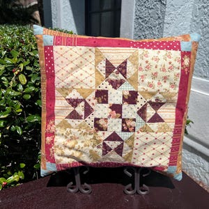 May include: A square decorative pillow with a patchwork design. The pillow features a central star pattern in shades of burgundy, cream, and gold, surrounded by a red border. The corners are accented with light blue fabric.
