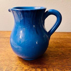 May include: A ceramic pitcher with a rounded body and a curved handle, featuring a vibrant blue glaze with darker blue vertical drip patterns running down the sides from the rim, and a small spout for pouring liquids.