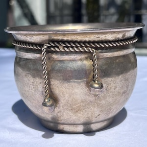 May include: A small, silver-toned bowl with a rounded body and a flared rim. The bowl is adorned with a twisted rope detail around the middle and two hanging rope accents with small ball-shaped ends. The bowl sits on a white surface.