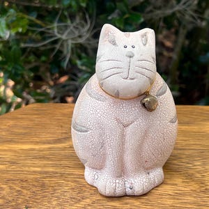 May include: A ceramic cat figurine in a seated position, with a cracked, off-white finish. The cat has a gold-colored bell on a gold-colored collar. Gray stripes accent the ears and sides. The figurine sits on a wooden surface.