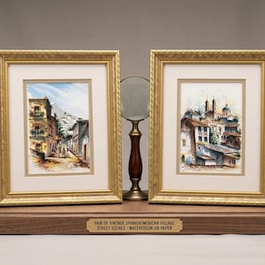 May include: Two framed watercolor paintings of Spanish/Mexican village street scenes. The paintings are in gold frames and rest on a wooden base. A magnifying glass with a wooden handle sits between the frames. The text on the base reads: "PAIR OF VINTAGE SPANISH/MEXICAN VILLAGE STREET SCENES - WATERCOLOR ON PAPER."
