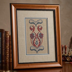 May include: Framed cross-stitch artwork featuring a symmetrical floral design in red, blue, and white. The artwork is set against a beige background, framed with a wooden frame and a black beaded inner border. The frame is displayed on a wooden surface.