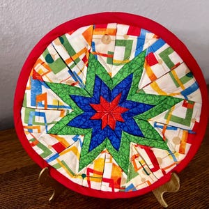 May include: A round, quilted trivet with a red border. The center features a starburst design in shades of blue, green, and red, surrounded by a patchwork of colorful geometric shapes. The trivet is displayed on a gold-colored stand.