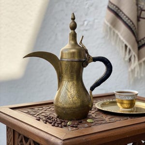 May include: A vintage brass coffee pot with an ornate design, a black handle, and a long spout. The pot sits on a carved wooden table with coffee beans and a small cup of coffee on a gold tray. A decorative cloth hangs in the background.