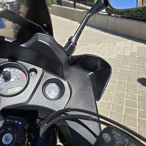 May include: A close-up shot of a black motorcycle dashboard featuring gauges, including a speedometer and fuel gauge. The motorcycle has a black windscreen and a side mirror reflecting the sky. Handlebars and controls are visible.