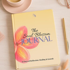 Op de afbeelding: Een gele journal met de titel "The Soul Blossom Journal" met een bloemmotief. De journal bevat de tekst "15 Days of Reflection, Healing & Growth". Een gouden pen, een kopje koffie en een bril liggen ook op tafel.