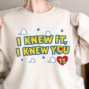 May include: A beige crewneck sweatshirt featuring the yellow text I KNEW IT, I KNEW YOU with a blue outline. The design includes a red heart with the letters TS, a ball with a red star, and small blue clouds and stars printed on the front.