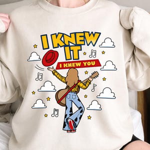 May include: A beige crewneck sweatshirt featuring a graphic illustration of a person with long hair playing a guitar, wearing a yellow plaid shirt and cow-print bell-bottom jeans. The text reads I KNEW IT and I KNEW YOU, with clouds, stars, and musical notes.