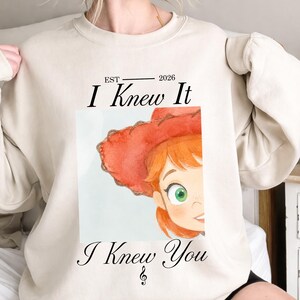 May include: A beige crewneck sweatshirt featuring a graphic print of a cartoon character with red hair and a red hat. The text reads EST 2026 I Knew It I Knew You with a small musical treble clef symbol centered below the text on the lower portion.