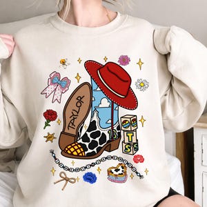 Puede incluir: Una sudadera beige de cuello redondo con un estampado gr&aacute;fico de una bota vaquera con manchas de vaca, un sombrero rojo, el nombre TAYLOR, el n&uacute;mero 13, bloques con las letras T, S, un lazo, flores, estrellas y una cadena de cuentas.