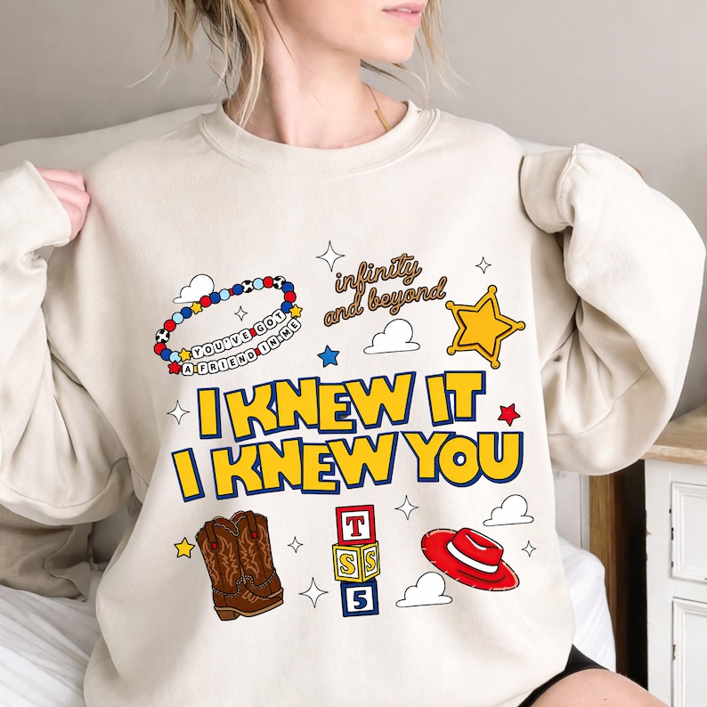 May include: A beige crewneck sweatshirt featuring a graphic print with the text "I KNEW IT I KNEW YOU" in yellow with blue outlines. The design includes a beaded bracelet, a sheriff star, cowboy boots, alphabet blocks, and a red hat.