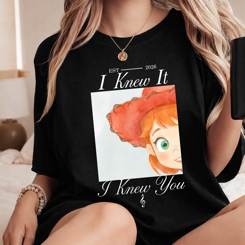May include: A black t-shirt featuring a white square graphic with a watercolor-style illustration of a character with red hair and a red hat. The text reads EST 2026, I Knew It, and I Knew You with a small musical note symbol below the text.
