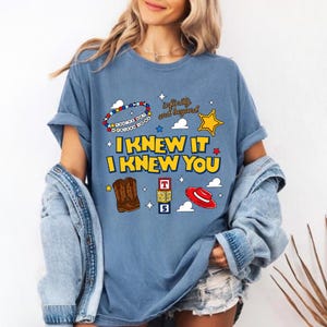 May include: A blue t-shirt featuring a graphic print with the text "I KNEW IT I KNEW YOU" in yellow, "infinity and beyond" in brown, and "YOU'VE GOT A FRIEND IN ME" on a beaded bracelet. Includes illustrations of brown cowboy boots, a red hat, and blocks.
