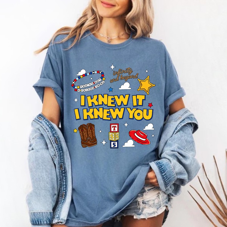 May include: A blue t-shirt featuring a graphic print with the text "I KNEW IT I KNEW YOU" in yellow, "infinity and beyond" in brown, and "YOU'VE GOT A FRIEND IN ME" on a beaded bracelet. Includes illustrations of brown cowboy boots, a red hat, and blocks.